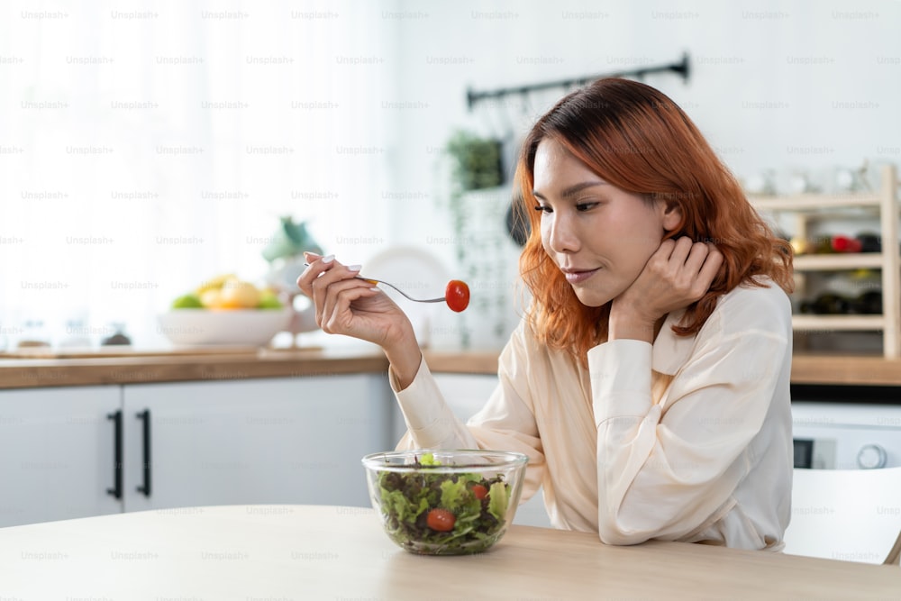 Asian young happy attractive woman eat green salad in kitchen at home. Beautiful girl feel joyful and enjoy eating vegetables healthy foods to diet and lose weight for health care wellness in house.