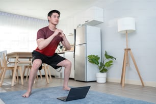 Asiatischer junger gutaussehender Mann, der Cardio-Übungen im Wohnzimmer zu Hause macht. Attraktiver aktiver Mann, der Hockentraining auf dem Boden durchführt, folgen Sie dem Anweisungsvideo vom Online-Trainer auf dem Laptop-Computer im Haus.