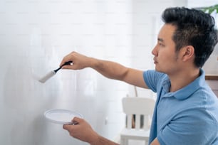 Asian attractive man paint white wall with brush to repair living room. Young handsome male painter use roller color to cover dirty wall for renovate and decorate room. Home renovation renewal concept