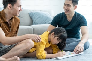 Asian attractive LGBTQ gay family teach young girl kid draw picture. Handsome male couple look at little adorable child daughter play and coloring book in living room, enjoy parenting activity at home