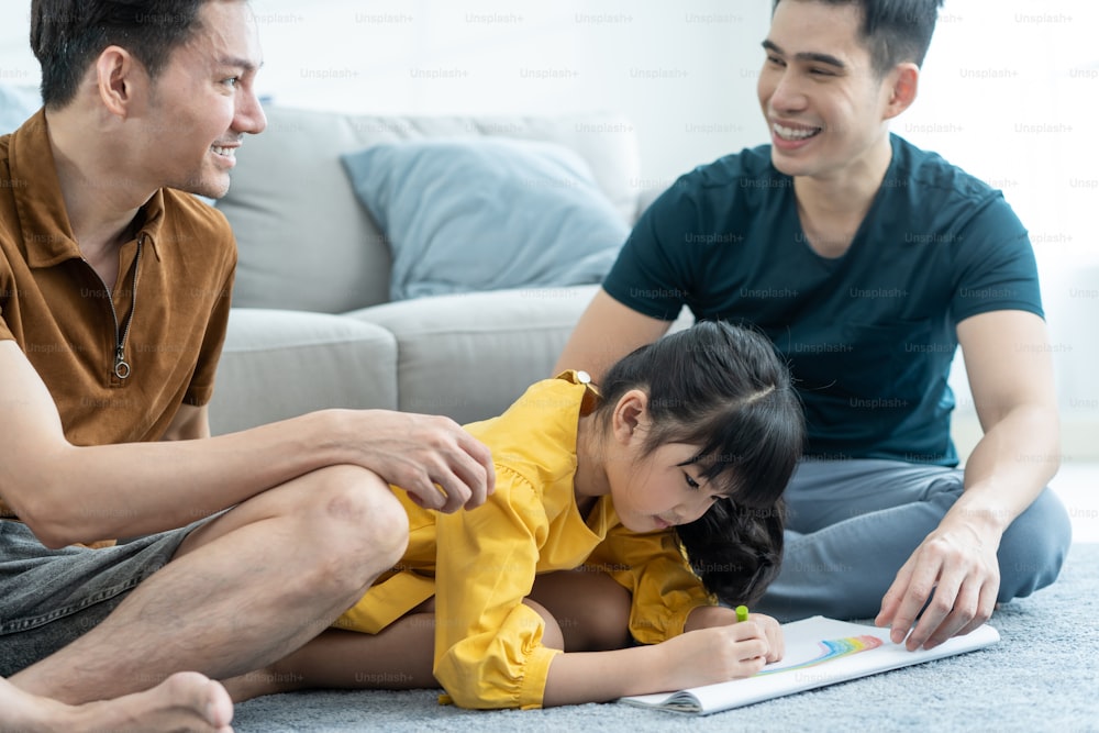 Asian attractive LGBTQ gay family teach young girl kid draw picture. Handsome male couple look at little adorable child daughter play and coloring book in living room, enjoy parenting activity at home