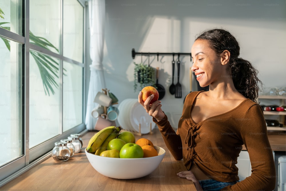 Young attractive Latino woman eat fruits on table in kitchen at home. Beautiful active female stand with happy face, enjoy eating banana, healthy foods for health care wellness to lose weight in house