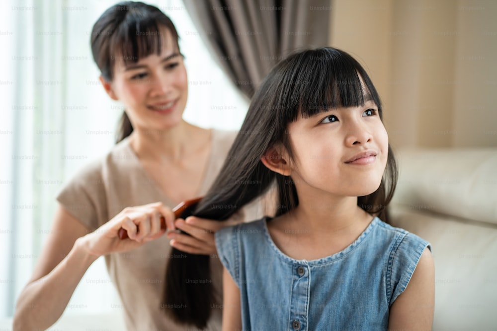 Asian loving mother spend leisure time with young little girl daughter in living room in house. Caring mom combing small child's hair with hairbrush with gentle. Family parenting relationship at home