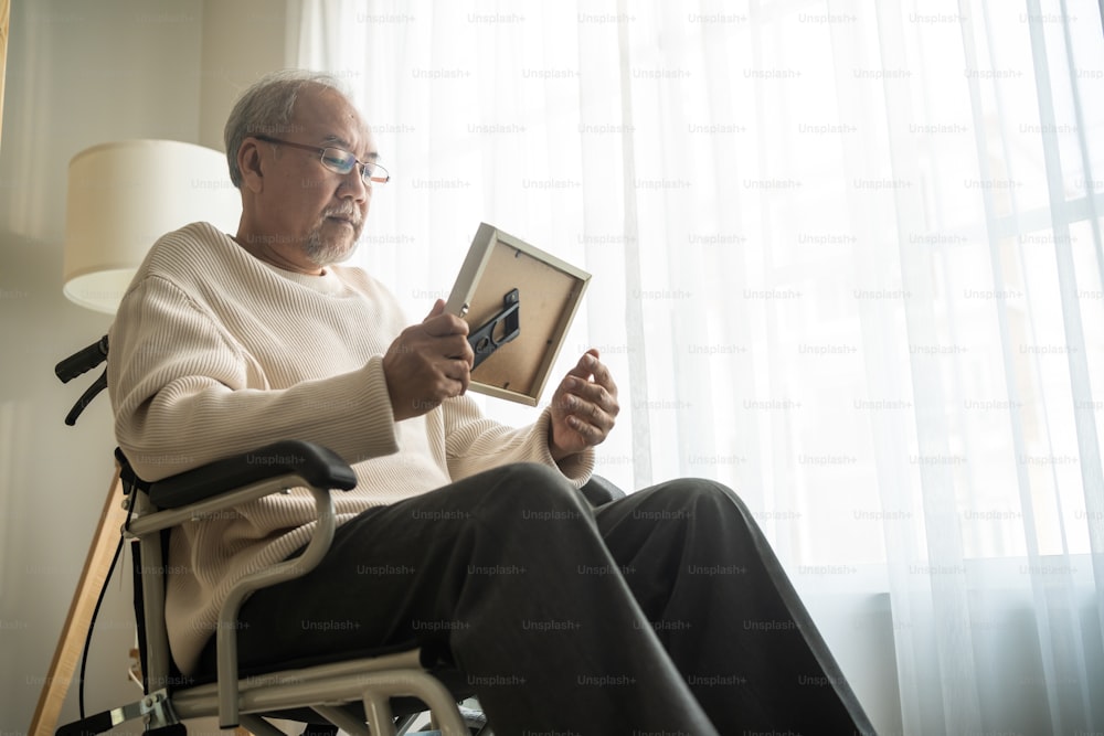 アジアの年配の男性は車椅子に一人で座り、家族写真を見ています。高齢の高齢の成熟した祖父は、退職後に老人ホームに滞在している間、親戚と妻を恋しく思い、孤独で悲しいと感じています。