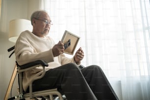 Un hombre mayor asiático se sienta solo en una silla de ruedas y mira fotografías familiares. El abuelo anciano y maduro se siente solo y triste, extrañando a su pariente y esposa mientras permanece en un hogar de ancianos después de la jubilación.
