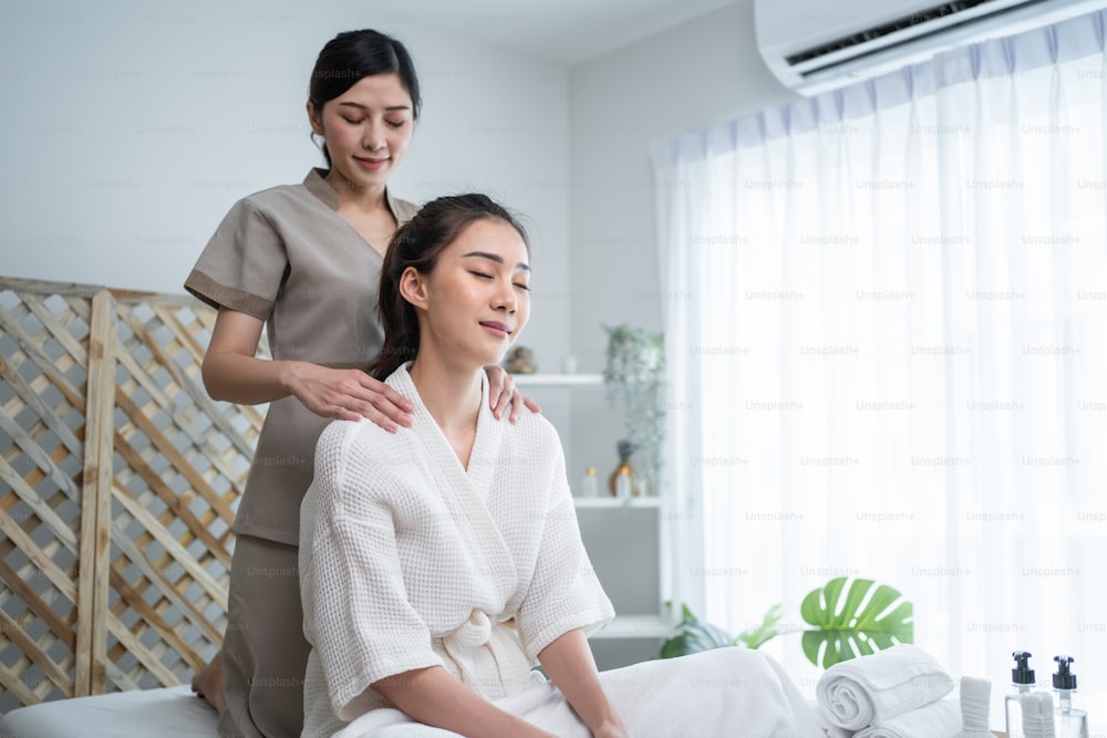 Asian young happy woman feeling relax during shoulder and back massage. Attractive beautiful girl sitting on massage table getting physiotherapy service from masseuse for body care in spa beauty salon