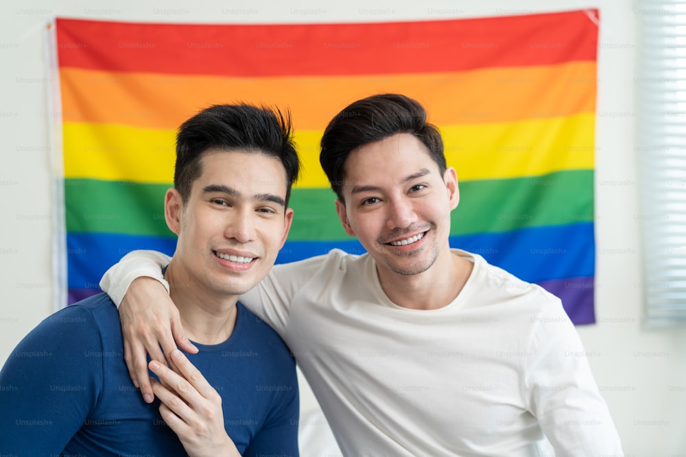 Portrait of Asian handsome man gay family holding LGBT flag and smile. Attractive romantic male lgbt couple sit on bed in bedroom in morning and looking at camera with gay pride and rainbow background