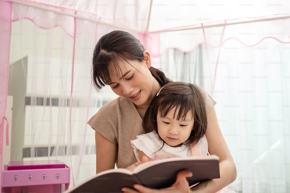 Una madre asiática leyó un cuento a su hija en una tienda de campaña en casa. La hermosa madre juega al libro ilustrado del juego de la educación con un niño lindo y feliz para desarrollar la habilidad. Concepto de actividad de crianza en casa