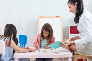 Caucasian beautiful young woman teacher teaching art to kids at school. Attractive Instructor master female explain and educate drawing and painting with student kindergarten pre school in classroom.