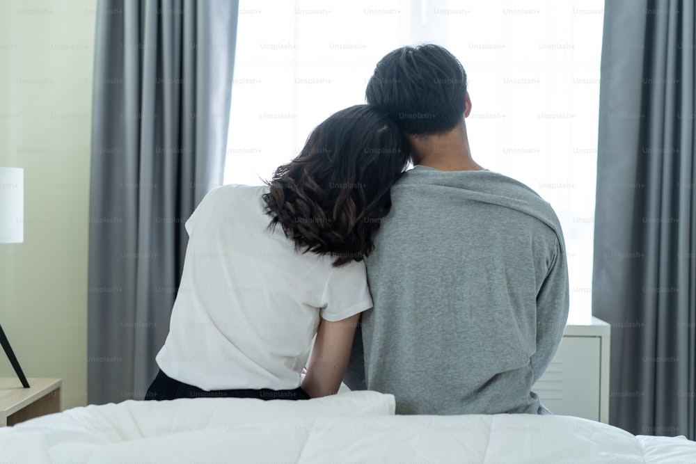 Rear view of Asian couple sitting together after wakeup in the morning. Attractive new marriage man and woman in pajamas feel happy and relax in the early day after get up from bed in bedroom together