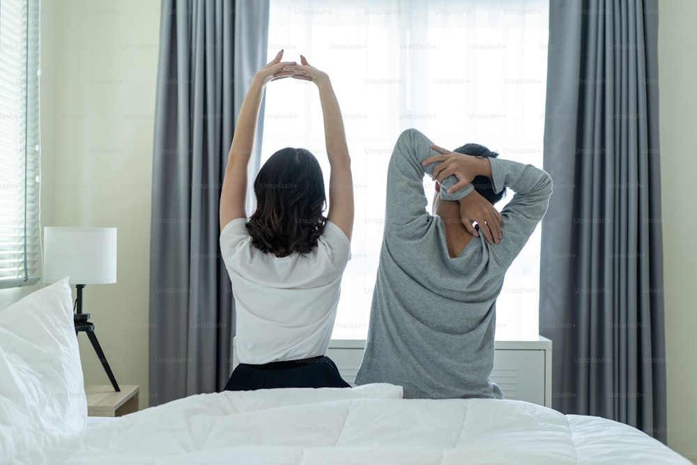 Rear view of Asian couple stretching hands after wakeup in morning. Attractive new marriage man and woman in pajamas feel happy and relax in the early day after get up from bed in bedroom together.