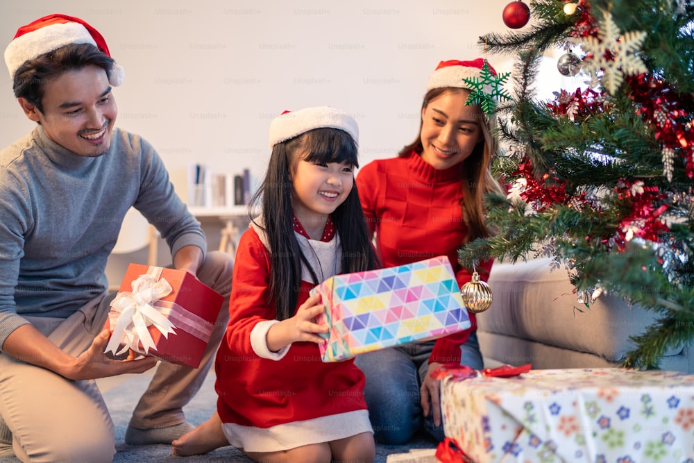 Adorabile membro della famiglia asiatica che decora l'albero di Natale con regalo regalo. Giovane figlia che si sente felice ed entusiasta di celebrare la festa del Ringraziamento di Natale durante le vacanze insieme ai genitori in casa.
