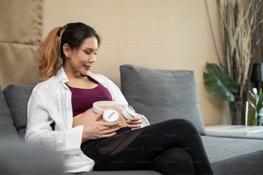 Asiatische junge Frau, schwanger sitzend und Kopfhörer auf dem Bauch haltend. Attraktive weibliche Schwangerschaft Sitzen Sie auf Komfort-Sofa Öffnung spielen Musik für Baby im Mutterleib mit Glück und lächeln im Wohnzimmer zu Hause