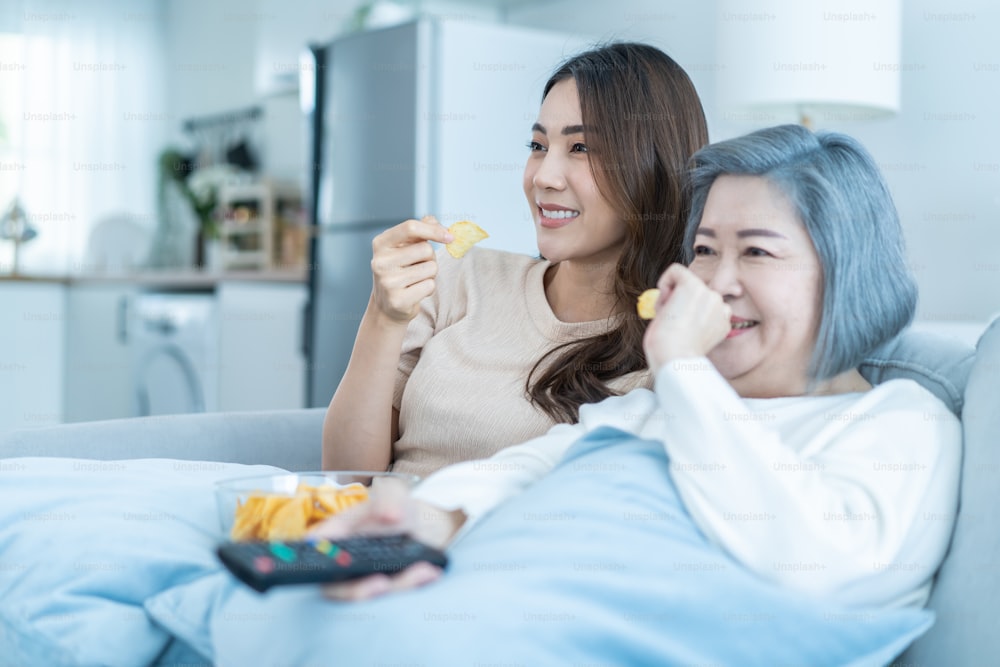 Asian senior mature woman and daughter sit on sofa watch funny movie. Loving elderly older mother spend time in living room at home with beautiful girl enjoy comedy show on TV and eat snacks together.
