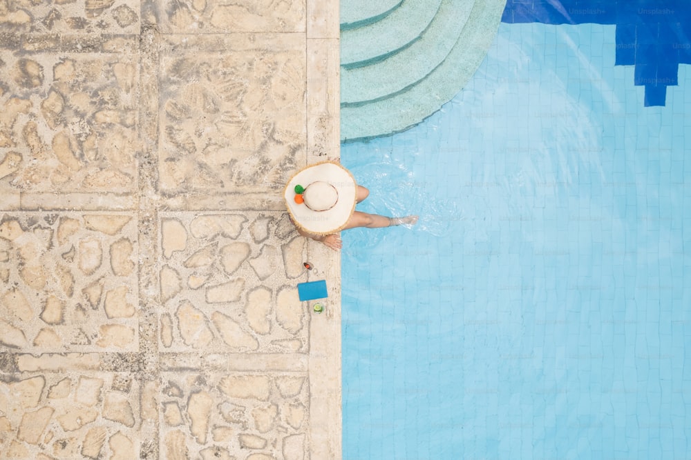 Vue aérienne d’une femme au chapeau de soleil se relaxant, prenant un bain de soleil au bord de la piscine d’été