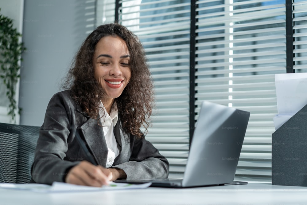 Happy beautiful Latino businesswoman in formal suit work in workplace. Attractive female employee office worker smile, use laptop computer for discuss with co worker and take note on paper in company.