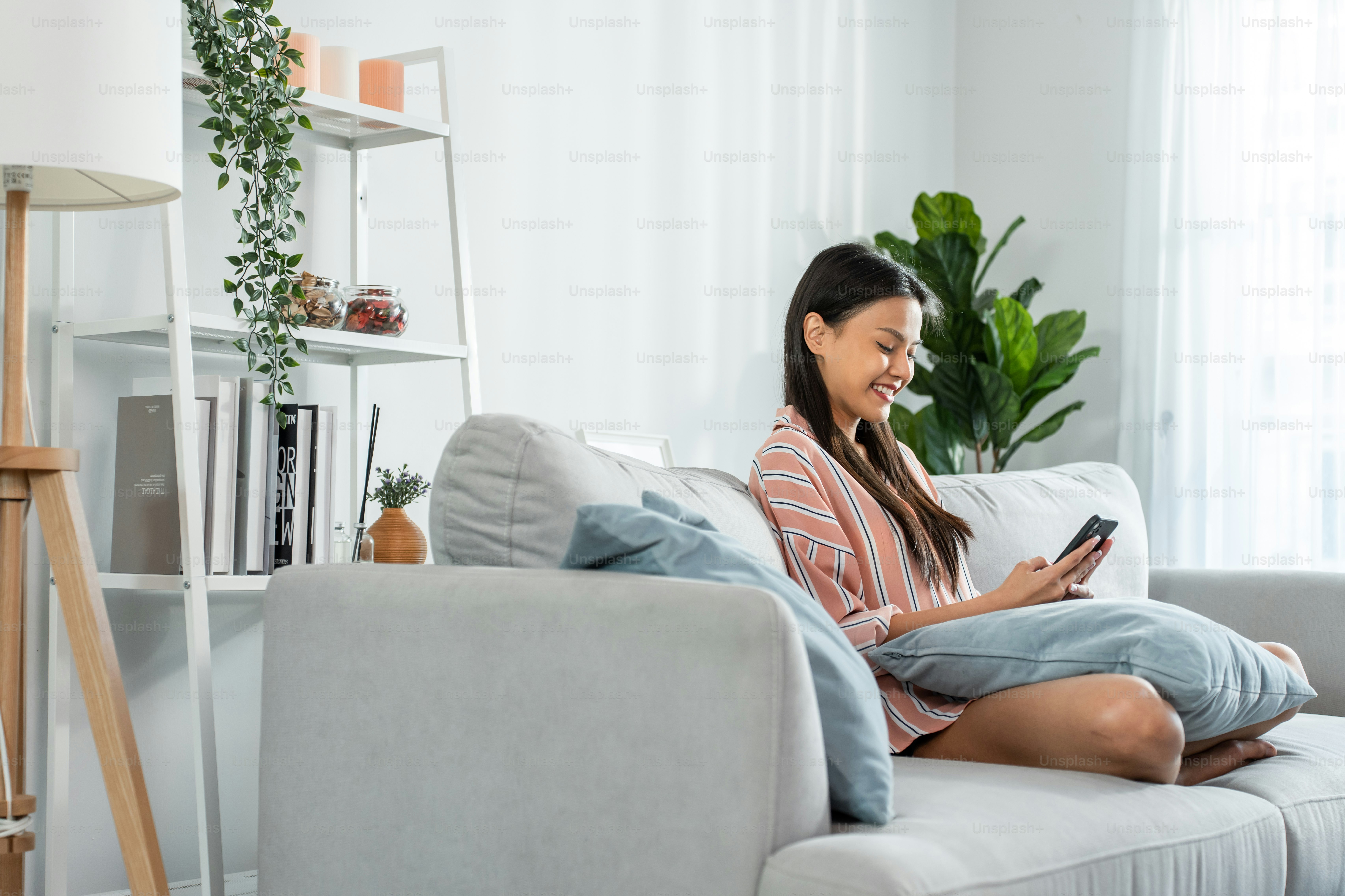 Belle femme asiatique assise sur un canapé et glissant le téléphone portable dans la maison. Heureuse jeune fille attrayante passer du temps libre à la maison, se sentir détendue et profiter de communiquer et discuter sur smartphone dans le salon