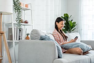 Asian beautiful woman sitting on sofa and swipe mobile phone in house. Happy attractive young girl spend leisure time at home, feel relax and enjoy communicate and discuss on smartphone in living room