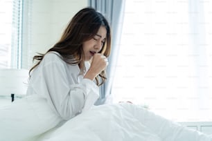 Asian beautiful sick girl in pajamas getting up from sleep in bedroom. Attractive young woman feeling bad after wake up on bed and coughing in early morning at home. health care-lifestyles concept.