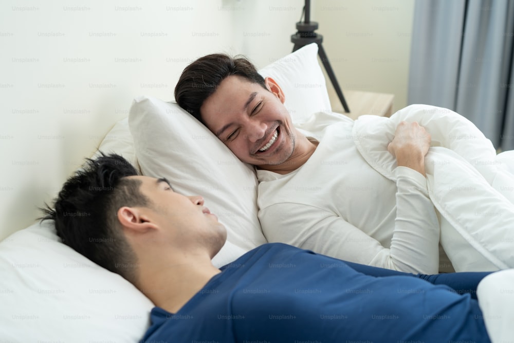 Asian handsome man gay couple lying down on bed and look at each other. Attractive romantic male lgbt in pajamas sleeping in early morning time together in bedroom at home. Homosexual-LGBTQ concept.