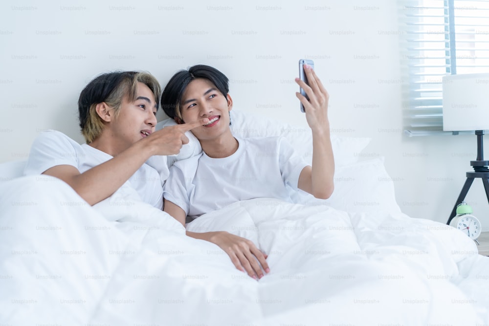 Asiático guapo hombre gay usar videollamada de teléfono móvil en línea con la familia. Atractivo guapo LGBTQ en pijama acostado en la cama en el dormitorio, sostenga el teléfono inteligente y hable de forma remota virtual por la mañana en la casa.