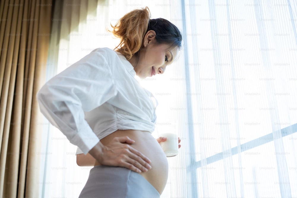 Asian beautiful young woman pregnant standing and drinking milk at home. Attractive female pregnancy stand and touching baby in the womb, drink milk healthy drink and feeling happiness in living room.