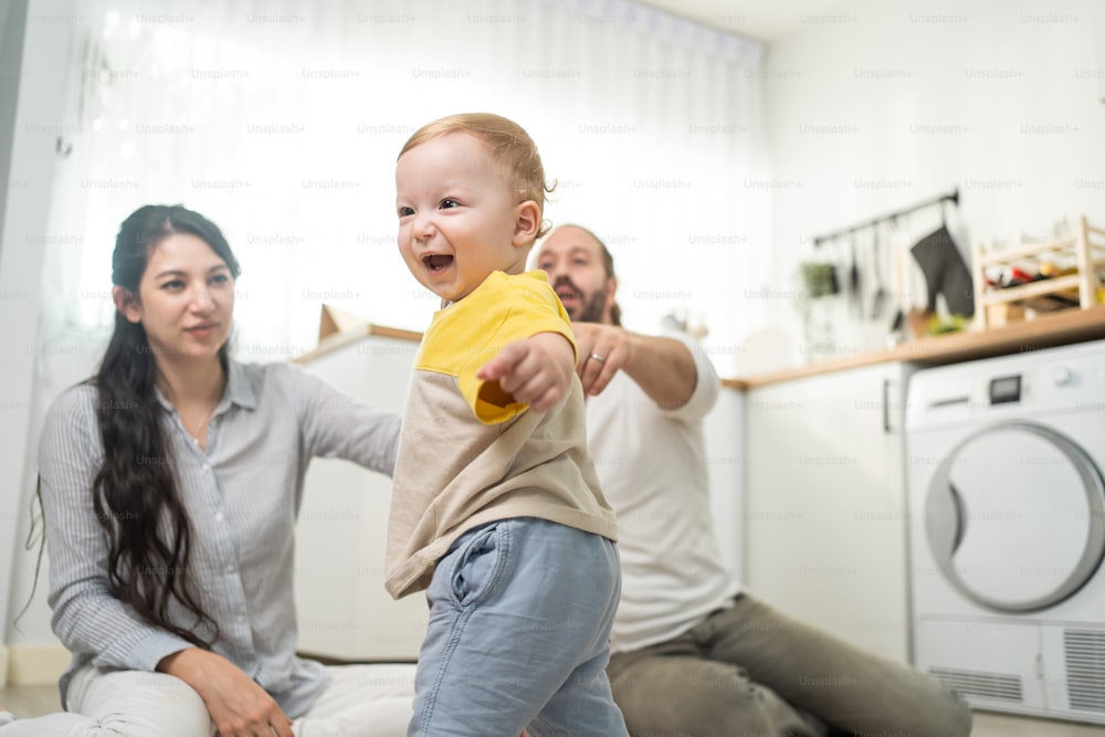 Caucasian baby boy child learn to walk with parents support in house. Happy family, mother and father helping young toddler son taking first step walk on floor to develop skill in living room at home.