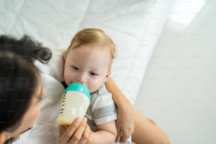 Caucasian beautiful mother holding and feeding baby from milk bottle. Attractive loving family, mom sit on bed and carry sleepy little infant toddler on hands and fed son drink from bottle in house.