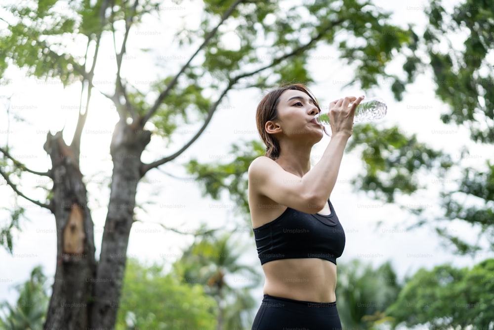 Atleta jovem asiática bebe garrafa de água após o exercício no parque. Esportista bonita atraente ativa fazer uma pausa após a ioga, correndo treino ao ar livre à noite para cuidados de saúde no jardim.