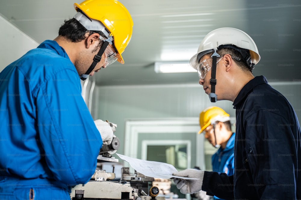 Trabalhadores mecânicos asiáticos trabalhando em uma fresadora. Os técnicos usam óculos de proteção e capacete ao operar a máquina por precaução de segurança. Líder aconselhando o membro de sua equipe a fazer um trabalho.