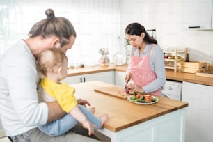 Caucasian beautiful parents cook food with baby boy toddler in kitchen. Happy family, Attractive young mother making healthy salad for lunch while husband father holding little kid son infant in house