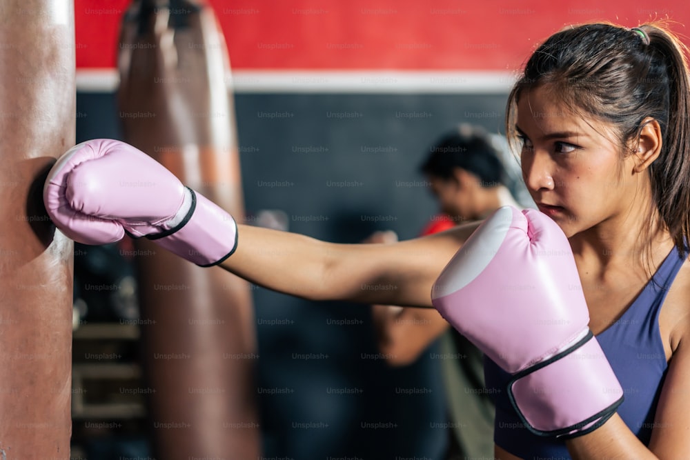 スポーツウェアとボクシンググローブを身に着けたアジアのアクティブな女性スポーツウーマン。フィットネスクラブで強い筋肉を維持するために砂袋を殴る練習をして運動する若い美しいアスリートの女の子。