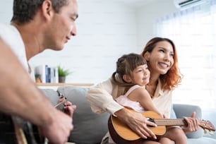 집에서 어린 아이와 함께 기타를 연주하는 아시아 젊은 부부. 매력적인 아름다운 부모님은 소파에 앉아 집안의 거실에서 노래를 부르는 어린 딸과 여가 시간을 보냅니다.