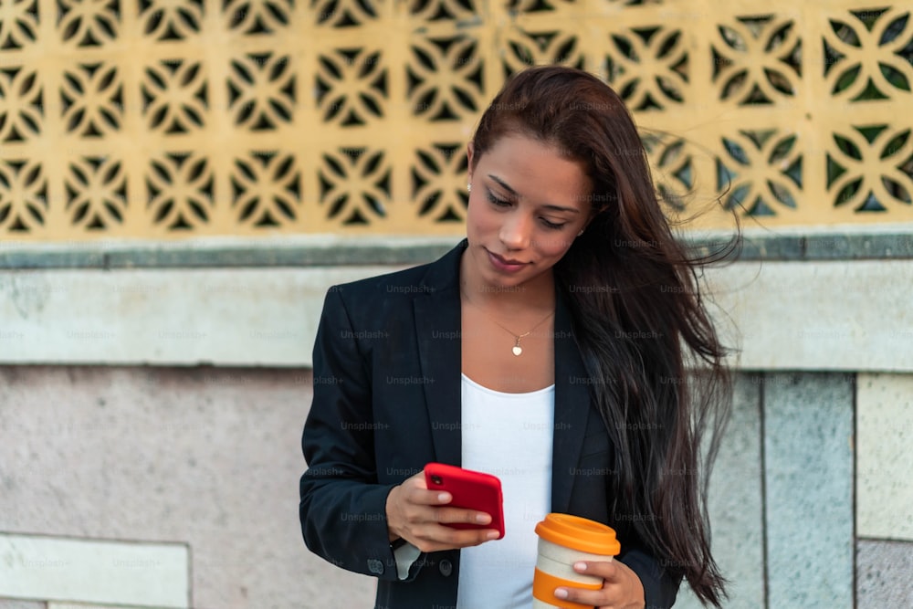 Junge Frau steht auf der Straße, trinkt Kaffee und benutzt ein Smartphone. Mädchen surft im Internet, chattet, bloggt. Frau hält das Telefon und schaut auf ihren Bildschirm.
