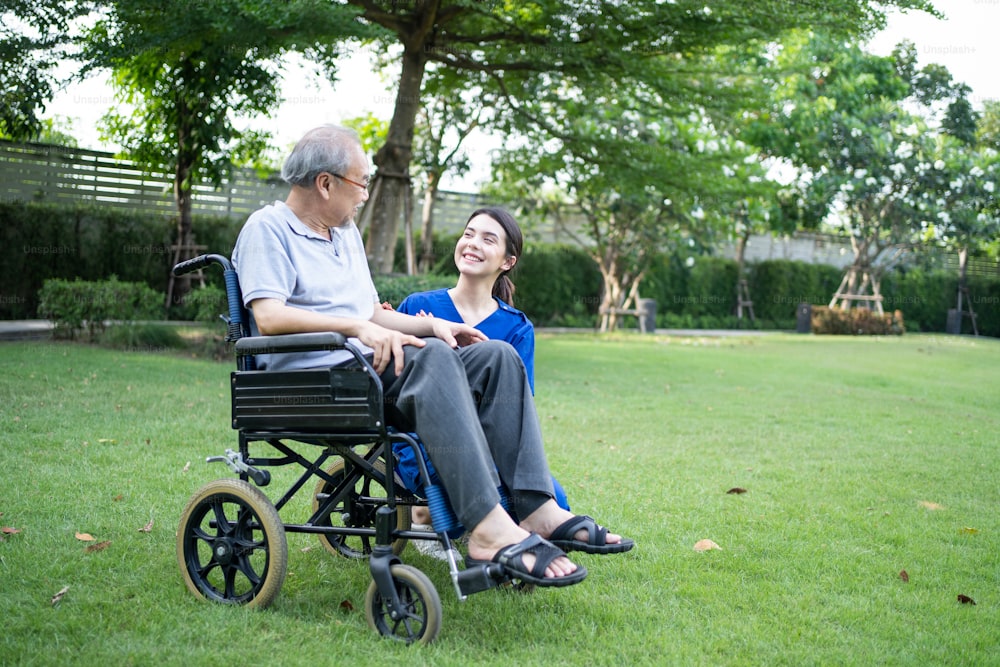 Asian young woman nurse at nursing home take care and support disabled senior elderly man on wheelchair at backyard. Beautiful Caregiver carer doctor and older patient enjoy talking and doing therapy.