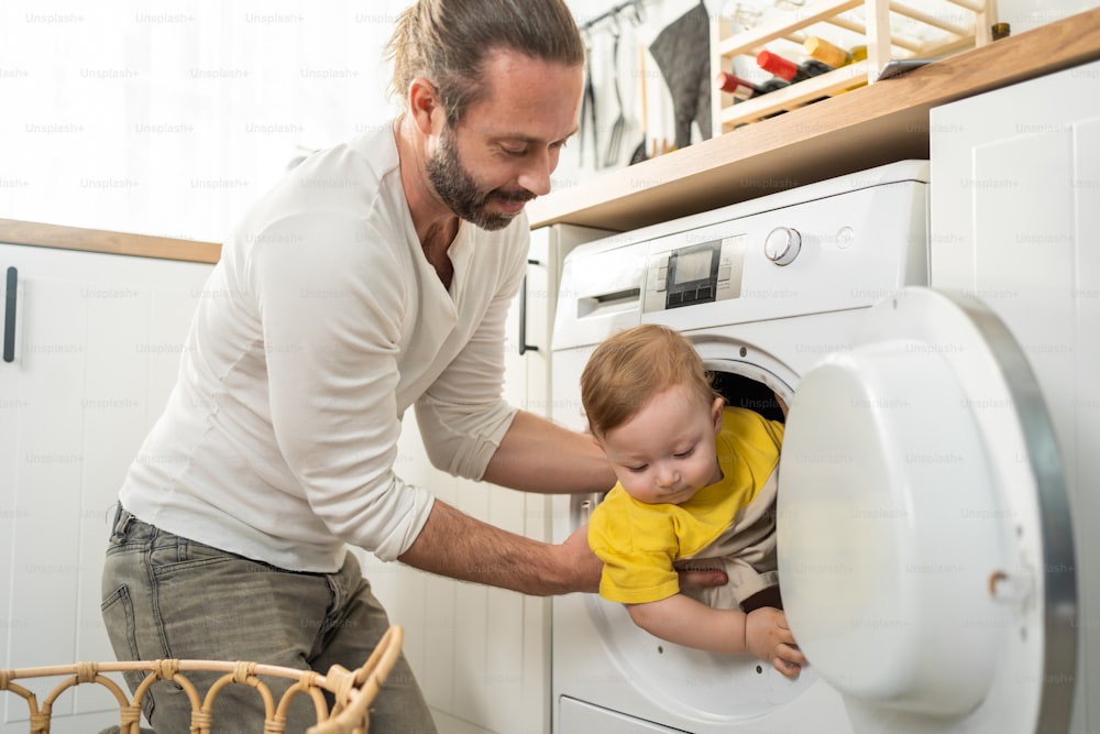 Caucasian loving parents play with baby toddler in kitchen on floor. Attractive young father spend free time and put naughty little son child in washing machine. Activity relationship at home concept.