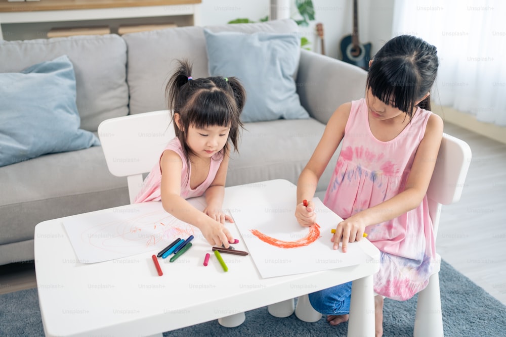 Asian young girl children drawing and coloring paper in living room. Little adorable sibling child sister sit on table, learn how to sketch art picture enjoy creativity activity on holiday at home.