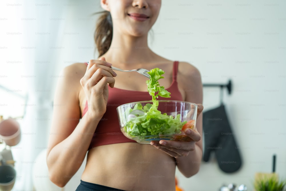 Close up da mulher asiática atraente segurar saladeira e comer vegetais. Menina do esporte bonito em roupas esportivas desfrutar de comer legumes limpos após o exercício para a saúde em casa. Conceito de Dieta e Alimentação Saudável.