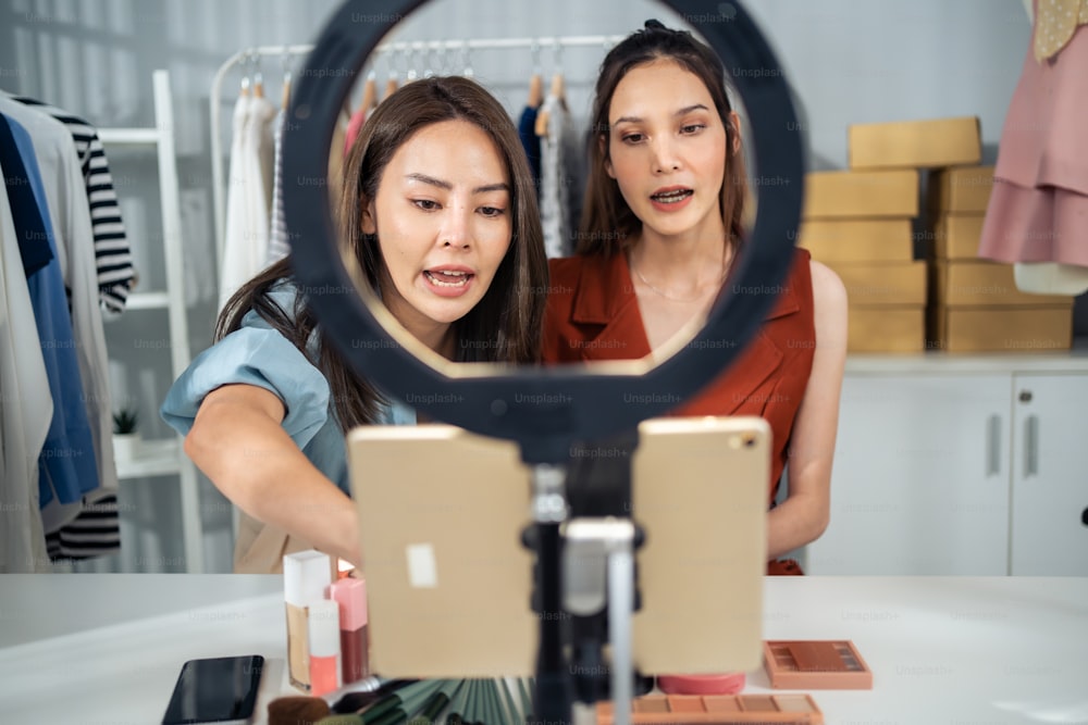 Dos vlogger asiáticas hablan a la cámara para revisar el cosmético en casa. Hermosa bloguera de belleza Influencer mujer amigas pareja transmitiendo en vivo y mostrando brocha de maquillaje a los seguidores en la tienda de la oficina.