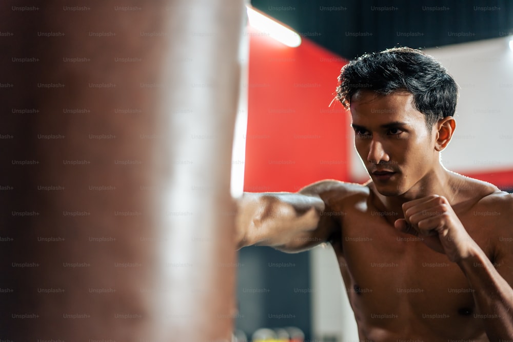 Asian active professional sport young man exercising in fitness gym. Attractive athlete sportsman workout by punching to sand bag to maintain strong muscle for health care in gym stadium or gymnasium.