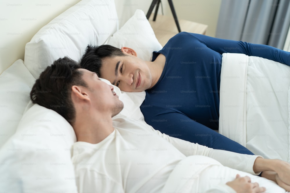 Asian handsome man gay couple lying down on bed and look at each other. Attractive romantic male lgbt in pajamas sleeping in early morning time together in bedroom at home. Homosexual-LGBTQ concept.