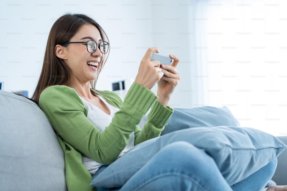 Asian woman wear eyeglasses play mobile game on smartphone at home. Attractive casual girl feel happy and relax, sit on sofa having fun touching screen on phone to play video enjoy victory in house.