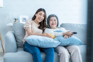 Asian senior mature woman and daughter sit on sofa watch funny movie. Loving elderly older mother spend time in living room at home with beautiful girl enjoy comedy show on TV and eat snacks together.