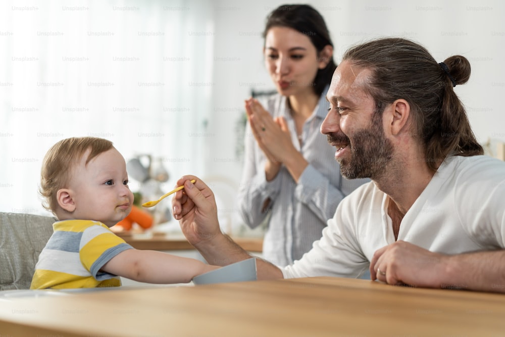 Caucasian young baby toddler eating healthy foods in kitchen at home. Little adorable cute kid boy infant sitting on children chair alone, enjoy having lunch in house. Child development skill concept.