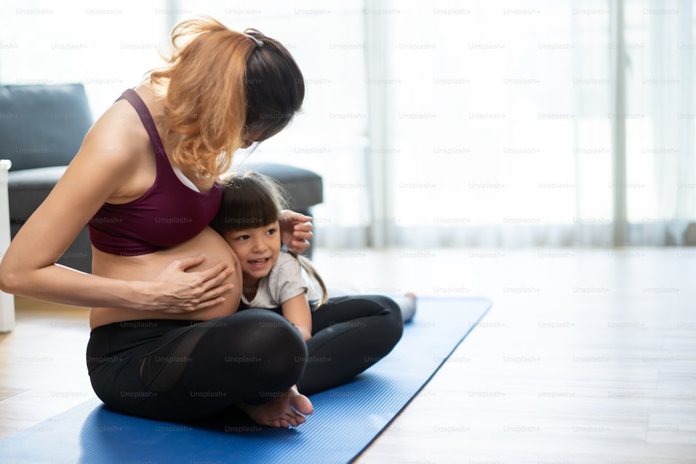 Asian young woman pregnant and little girl doing Yoga Pilates at home. Attractive mother pregnancy with kid daughter in sportswear feel happy and relax, follow exercise instruction from online trainer