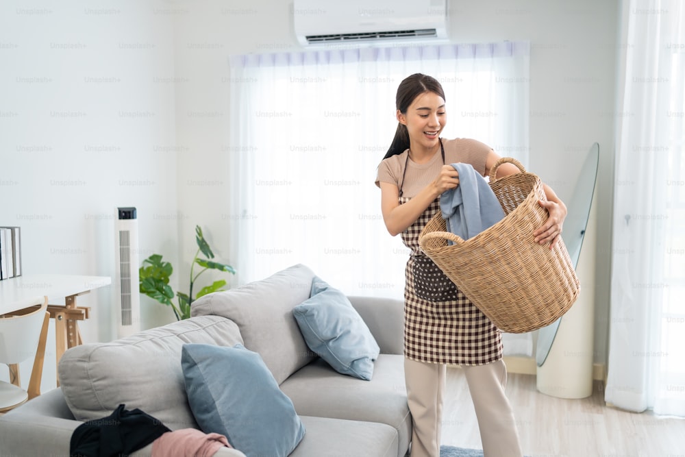 Asiatische Reinigungsdienstmitarbeiterin putzt im Wohnzimmer zu Hause. Schöne junge Mädchen Haushälterin Putzfrau fühlen sich glücklich und nehmen unordentliche schmutzige Kleidung in den Korb für Hausarbeit oder Hausarbeiten.