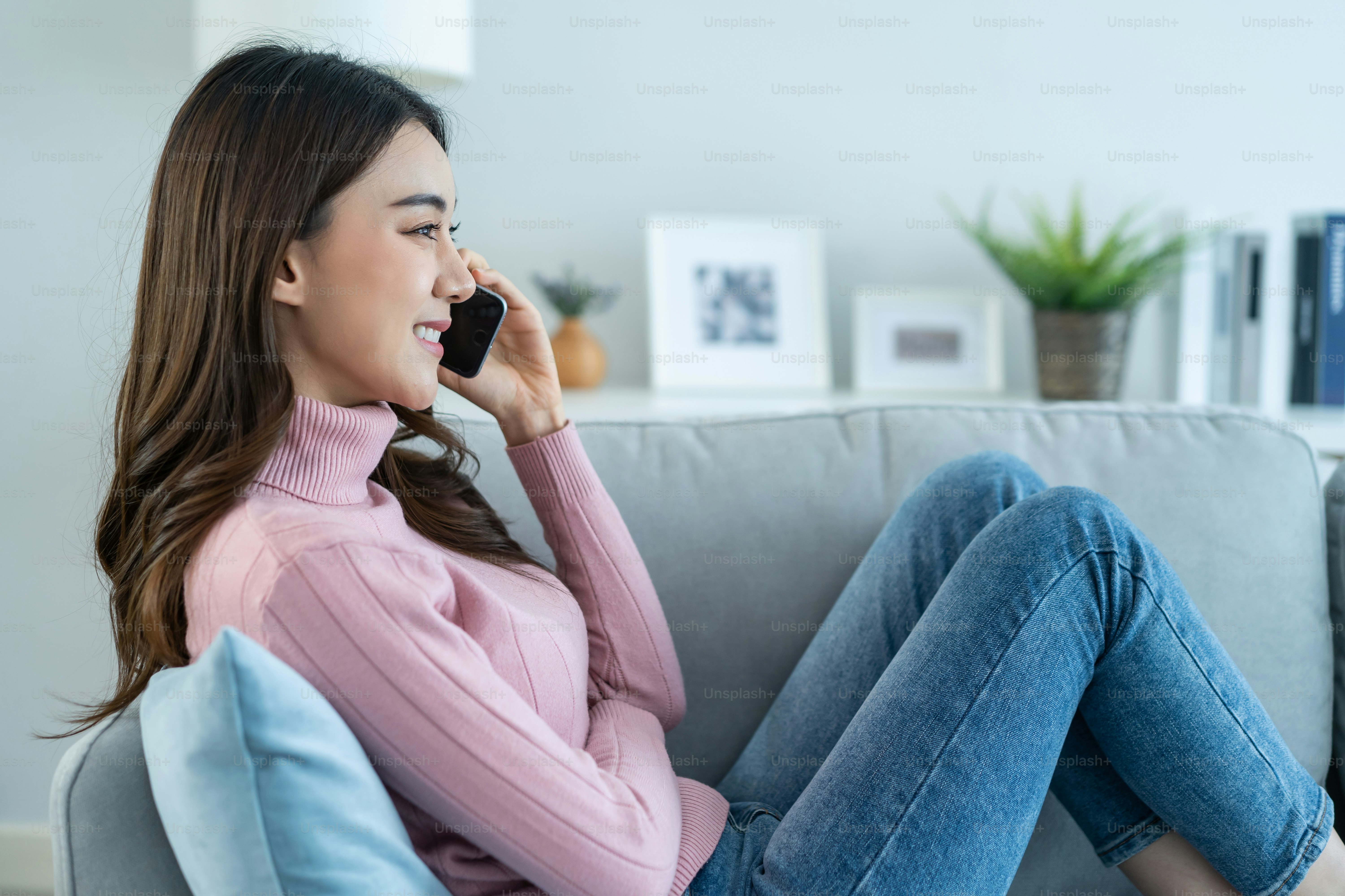 La bella donna asiatica si siede sul divano e parla al telefono cellulare in casa. La giovane ragazza attraente felice trascorre il tempo libero a casa, si rilassa e si diverte a comunicare e discutere sullo smartphone in salotto.