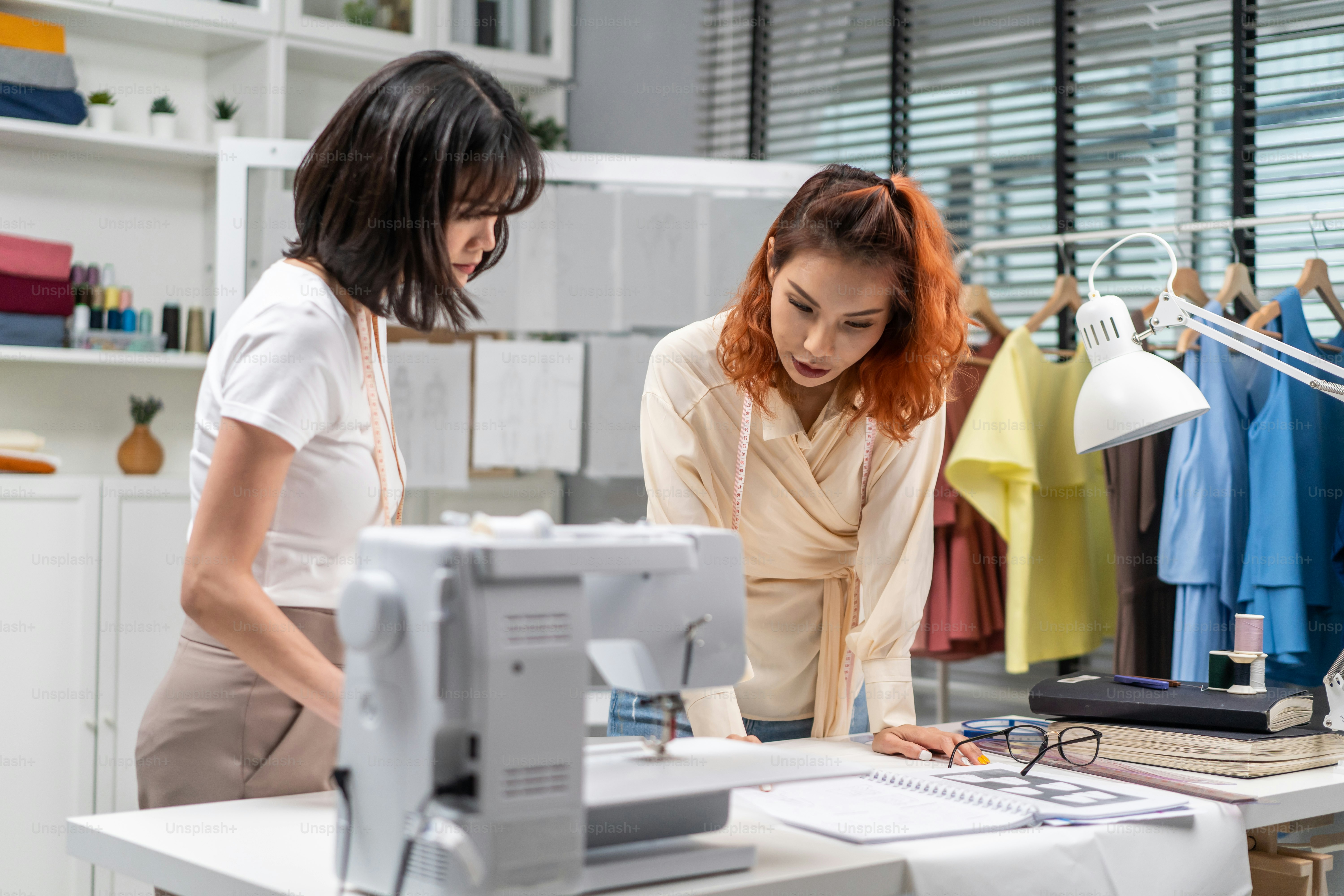 Asian two tailor woman working to design clothes in tailoring atelier. Attractive young female fashion designer dressmaker brainstorm to create new fashion pattern collection together in workshop room