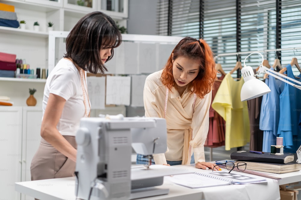 Asiatische zwei Schneiderinnen, die im Schneideratelier Kleidung entwerfen. Attraktive junge Modedesignerin Brainstorming, um gemeinsam im Workshop-Raum eine neue Modemusterkollektion zu kreieren
