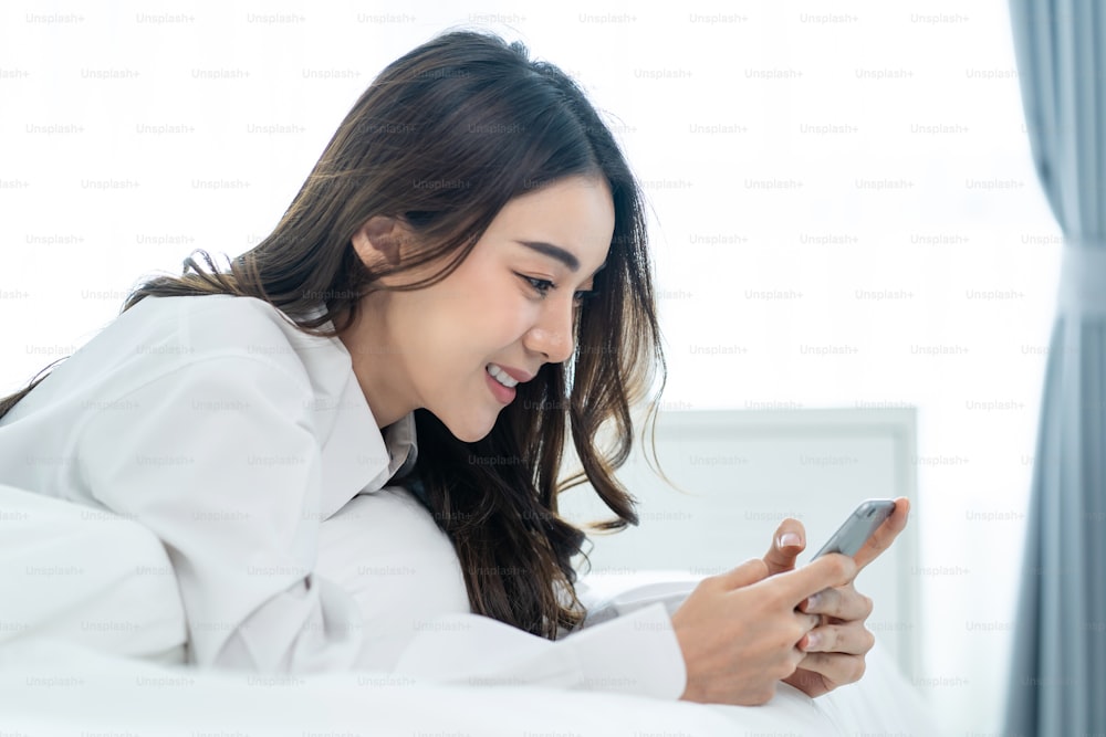 Asian attractive woman use mobile phone chat on bed at home in morning. Happy casual beautiful female lying down in bedroom enjoy holiday weekend, use smartphone communicate with technology in house.